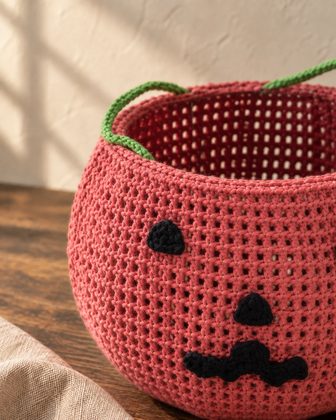 Close view of the pink crochet pumpkin-face basket with its green handle and black stitched features.
