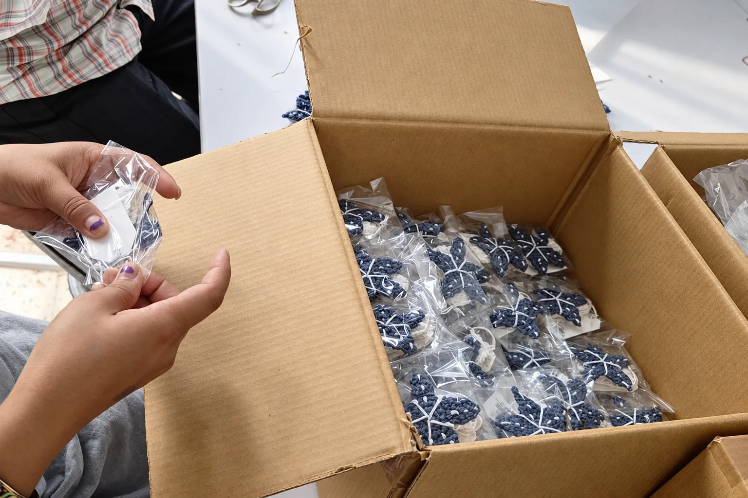 Hands packing individually wrapped crochet items into a cardboard box.