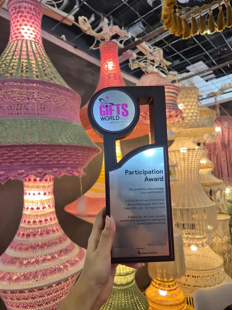 A hand holding our Participation Award from Gifts World Expo 2026, held at Bharat Mandapam, New Delhi, from 30 July to 1 August 2026, in front of lit crochet lamps.