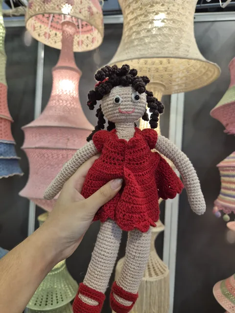A crochet doll in a red dress and red shoes, held up in front of pink, cream and green crochet lamps at our stall, Bharatiya Vyapar Mahotsav 2026.