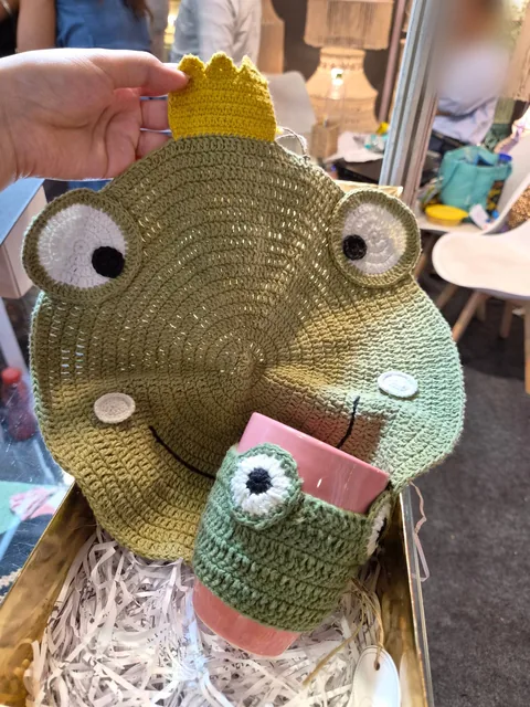A crochet frog-face mat with a yellow crown, held up above a pink mug in a matching crochet frog cosy, in a gift box at our stall, Gifts World Expo 2026.