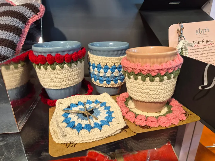 Mugs in crochet cosies with tulip and striped rims, on crochet coasters, beside a Glyph by Vera Verve thank-you card, at our stall, Gifts World Expo 2026.