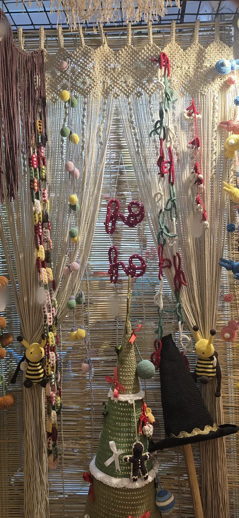 A cream macramé curtain hung with crochet strands, red hearts, bees and a small crochet Christmas tree, at our stall, IHGF Delhi Fair Spring 2026.