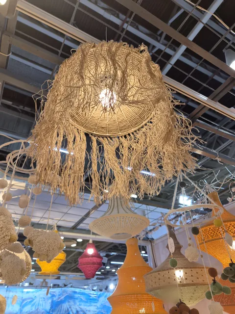 A fringed raffia pendant lamp lit from inside, hanging above crochet animal mobiles and orange, pink and yellow crochet lamps at our stall, IHGF Delhi Fair Autumn 2026.