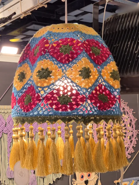 A crochet lampshade of red, yellow and blue granny squares with a fringe of golden tassels, lit at our stall, Bharatiya Vyapar Mahotsav 2026.
