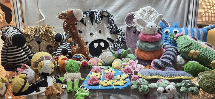 Crochet toys and cushions on a glass shelf at our stall, IHGF Delhi Fair Autumn 2025: zebras, a giraffe, a lion, frogs, turtles and a stacking ring toy.