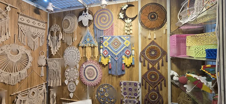 A corner of our stall at IHGF Delhi Fair Spring 2025 covered in macramé and crochet wall hangings and dream catchers, with pink, yellow and blue macramé baskets on glass shelves.