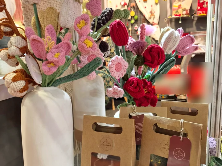 Crochet tulips, roses and wildflowers in white vases at our stall, beside Vera Verve gift bags holding crochet coasters.