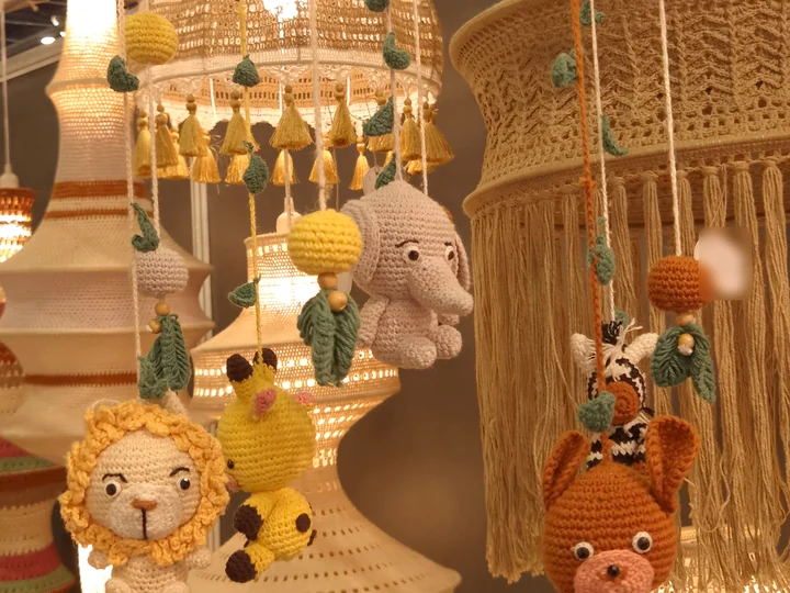 A crochet nursery mobile with a lion, giraffe, elephant, zebra and bear, hanging among lit lamps at our stall at Bharatiya Vyapar Mahotsav 2026.