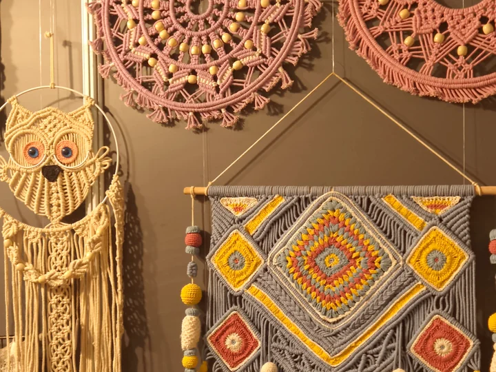 Macramé wall hangings on our stall wall at Bharatiya Vyapar Mahotsav 2026: a cream owl, pink mandalas and a blue, yellow and red diamond hanging.