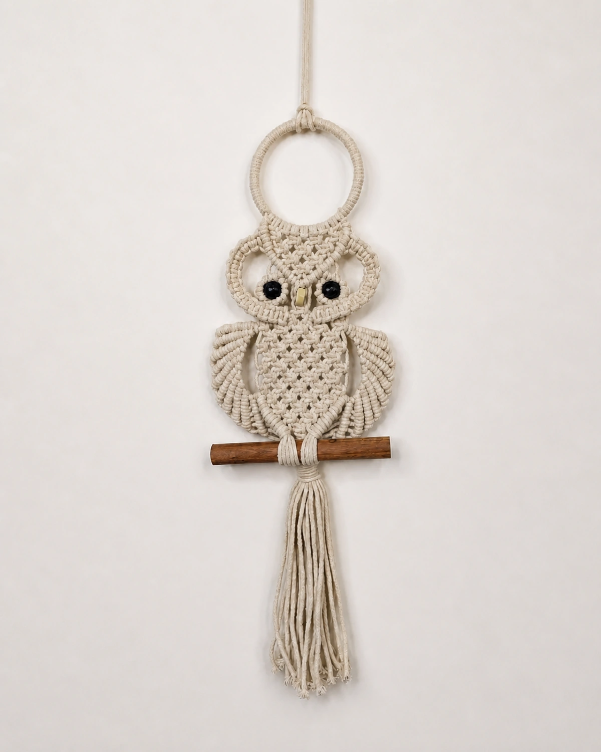 Cream macramé owl wall hanging perched on a wooden bar.
